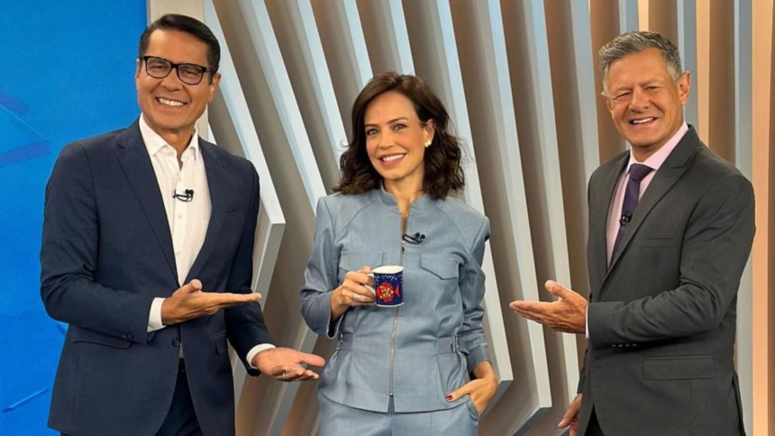 Os apresentadores Willian Leite (terno azul-marinho e óculos), Fabiana Oliveira (terninho cinza-azulado, segurando uma caneca) e Diógenes Lucca (terno cinza, gravata roxa) sorriem e posam no estúdio do Balanço Geral Manhã.