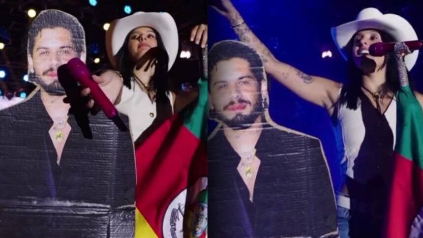 Cantora Ana Castela no palco com chapéu de cowboy e bandeira, ao lado de um boneco de papelão do cantor Zé Felipe, em show