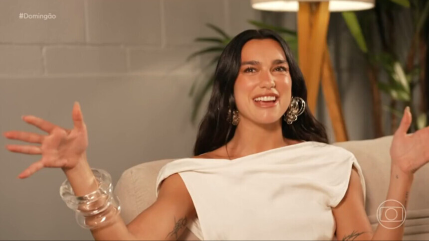 Cantora internacional Dua Lipa sorrindo e gesticulando durante entrevista para o programa Domingão com Huck