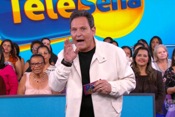 Luiz Ricardo sorridente e em pé, segurando um bilhete, no centro do cenário da Tele Sena Semanal edição 76
