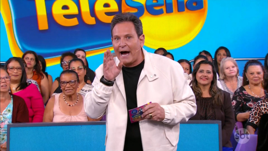 Luiz Ricardo sorridente e em pé, segurando um bilhete, no centro do cenário da Tele Sena Semanal edição 76