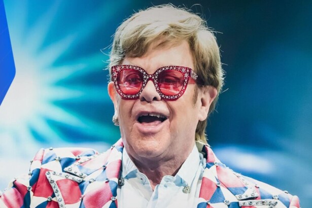 Sir Elton John em close-up, vestindo óculos de sol vermelhos e chamativos e casaco estampado, em apresentação ao vivo