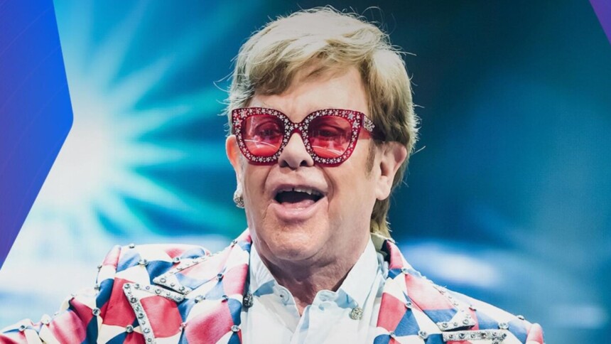 Sir Elton John em close-up, vestindo óculos de sol vermelhos e chamativos e casaco estampado, em apresentação ao vivo