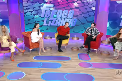 Apresentadores do Fofocalizando no SBT sentados em suas poltronas num cenário com cores predominantemente em tons de lilás e azul