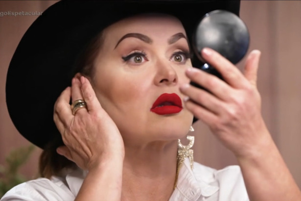 Gaby Spanic olha para o espelho em gravação para a RECORD. Ela usa chapéu de fazendeiro preto e batom vermelho escuro