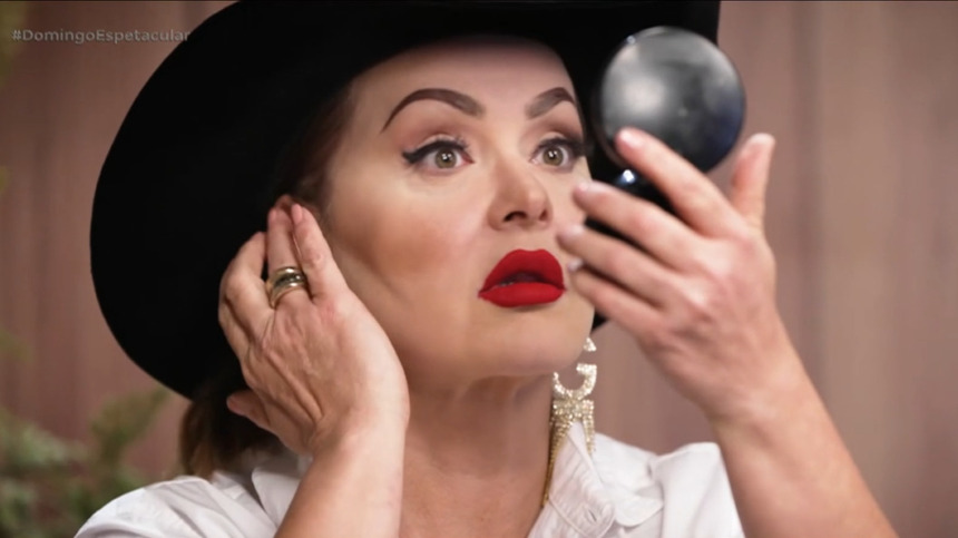 Gaby Spanic olha para o espelho em gravação para a RECORD. Ela usa chapéu de fazendeiro preto e batom vermelho escuro