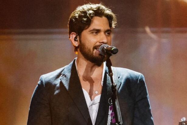 Cantor sertanejo Gustavo Mioto performando no palco, cantando em microfone com iluminação quente e vestindo terno e camisa branca