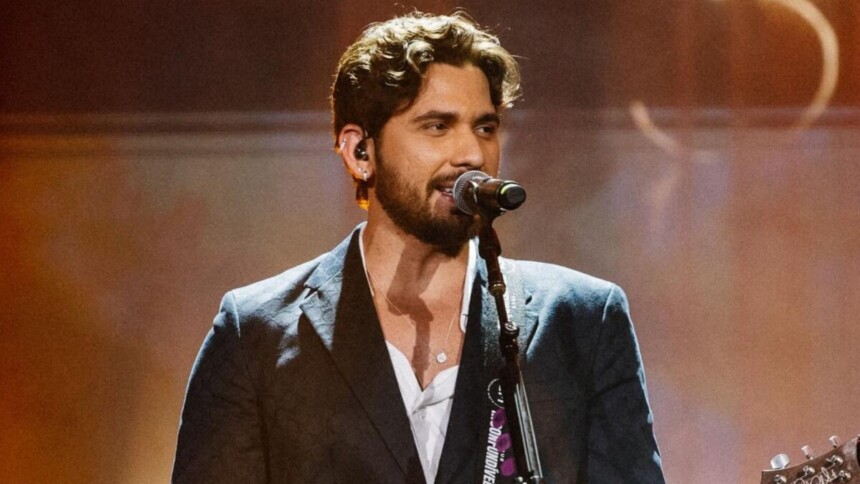 Cantor sertanejo Gustavo Mioto performando no palco, cantando em microfone com iluminação quente e vestindo terno e camisa branca