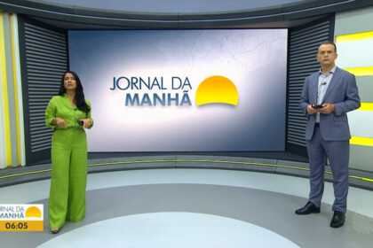 Camila Oliveira e Ricardo Ishmael em frente ao cenário do Jornal da Manhã com o grande telão atrás mostrando o logo do telejornal da afiliada da TV Globo