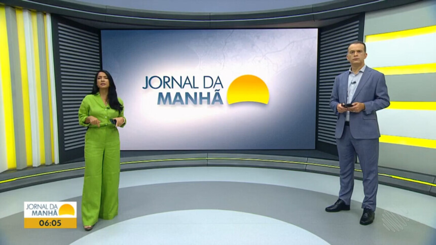 Camila Oliveira e Ricardo Ishmael em frente ao cenário do Jornal da Manhã com o grande telão atrás mostrando o logo do telejornal da afiliada da TV Globo