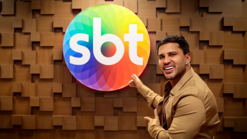 Lucas Guimarães apresentador do Eita, Lucas!, apontando e sorrindo ao lado do logo do SBT em comemoração à renovação de contrato