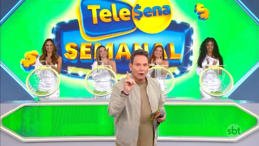 Luís Ricardo aponta para o alto e segura um bilhete da Tele Sena Semanal 77. O cenário está verde
