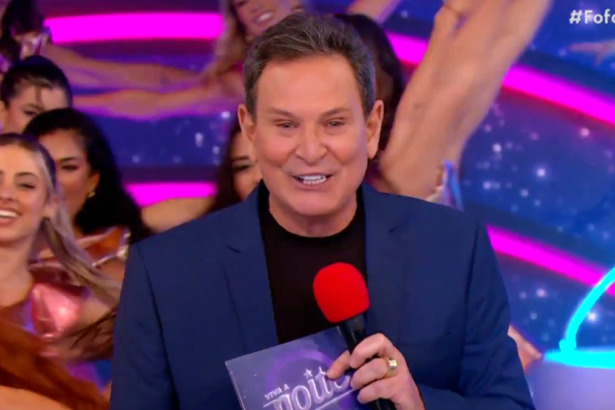 Luís Ricardo sorridente, com microfone e fichas na mão, no cenário do Viva a Noite