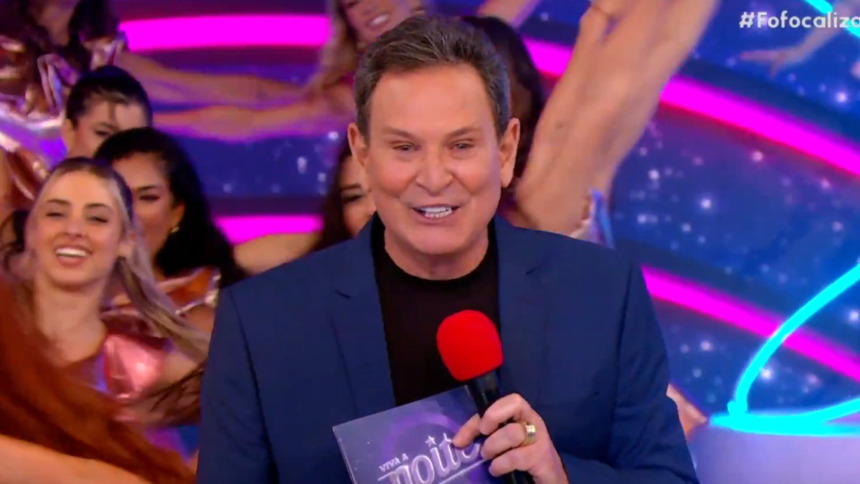 Luís Ricardo sorridente, com microfone e fichas na mão, no cenário do Viva a Noite
