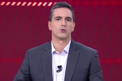 Jornalista Marco Pagetti apresentador do Aqui Agora do SBT, vestindo terno cinza em estúdio com fundo vermelho e luzes vibrantes