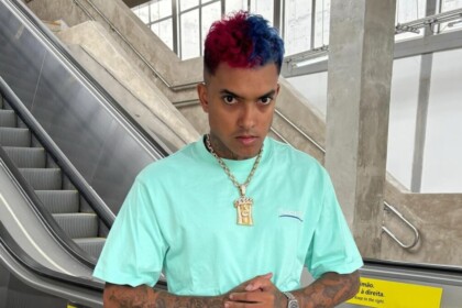 Cantor MC Brinquedo com cabelo metade azul e metade vermelho, vestindo camiseta azul-claro e corrente dourada, em frente a uma escada rolante