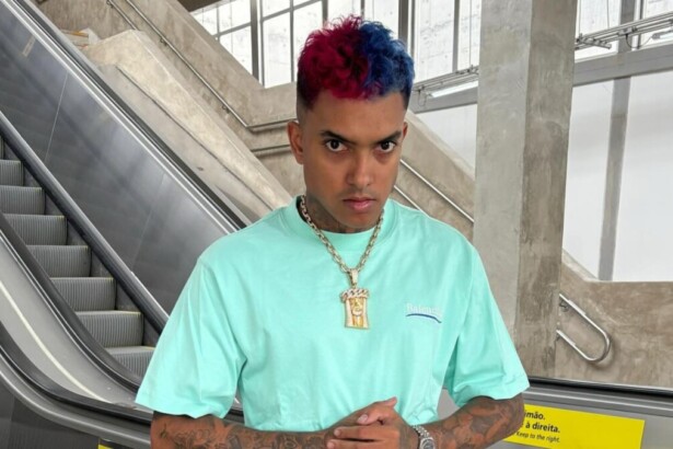 Cantor MC Brinquedo com cabelo metade azul e metade vermelho, vestindo camiseta azul-claro e corrente dourada, em frente a uma escada rolante