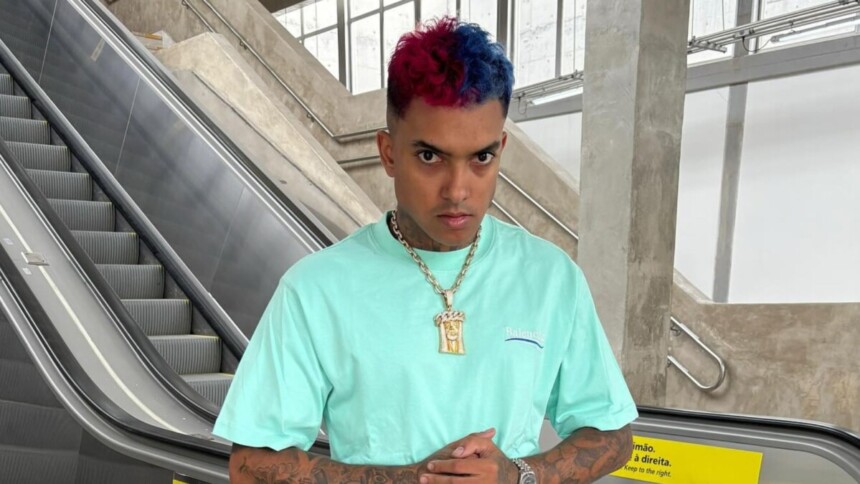 Cantor MC Brinquedo com cabelo metade azul e metade vermelho, vestindo camiseta azul-claro e corrente dourada, em frente a uma escada rolante