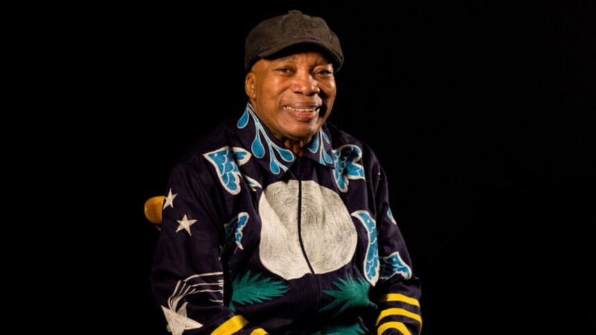 Cantor Milton Nascimento em retrato, sorrindo e vestindo jaqueta estampada com a lua e estrelas, em frente a um fundo preto