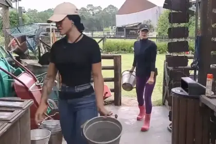 Rayane caminha na frente de Maria dentro da oficina de A Fazenda 17. Ela segura o balde de leite da vaca em uma das mãos