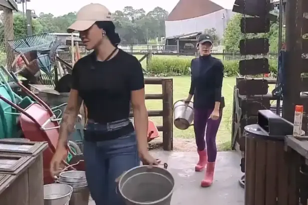 Rayane caminha na frente de Maria dentro da oficina de A Fazenda 17. Ela segura o balde de leite da vaca em uma das mãos