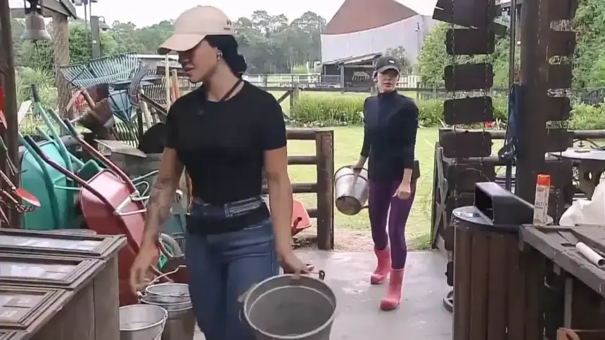Rayane caminha na frente de Maria dentro da oficina de A Fazenda 17. Ela segura o balde de leite da vaca em uma das mãos