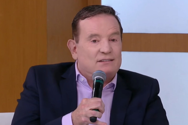 Roberto Cabrini segurando um microfone durante entrevista no programa Superpop, vestindo terno azul-marinho