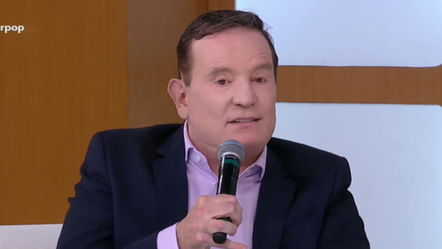 Roberto Cabrini segurando um microfone durante entrevista no programa Superpop, vestindo terno azul-marinho