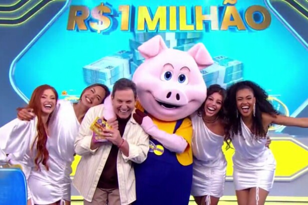 Luís Ricardo é abraçado pelo porquinho e pelas assistentes de palco da Tele Sena Semanal 80. No telão a frase "1 Milhão" sobre o prêmio da edição