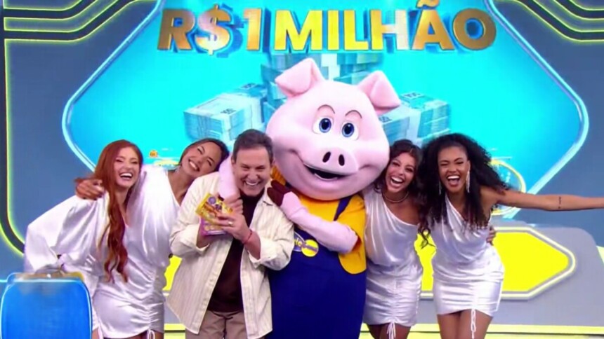Luís Ricardo é abraçado pelo porquinho e pelas assistentes de palco da Tele Sena Semanal 80. No telão a frase "1 Milhão" sobre o prêmio da edição