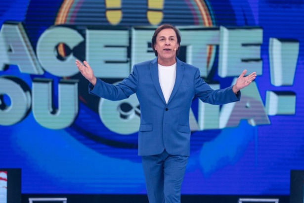 Tom Cavalcante apresentador do game show Acerte ou Caia, vestindo terno azul em cenário vibrante com o logo do programa