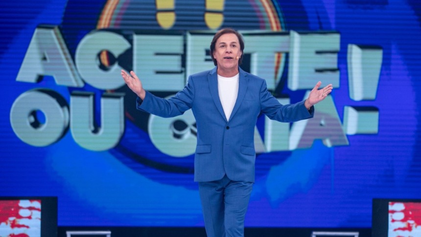 Tom Cavalcante apresentador do game show Acerte ou Caia, vestindo terno azul em cenário vibrante com o logo do programa