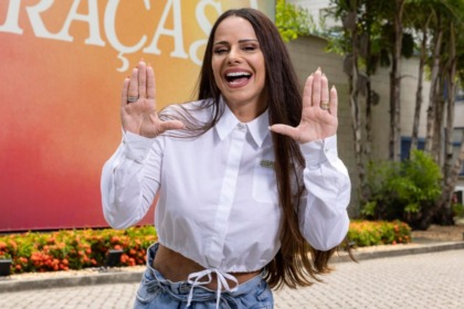 Atriz Viviane Araújo sorrindo e gesticulando em frente a um banner com o título da novela Três Graças, vestindo camisa branca e jeans