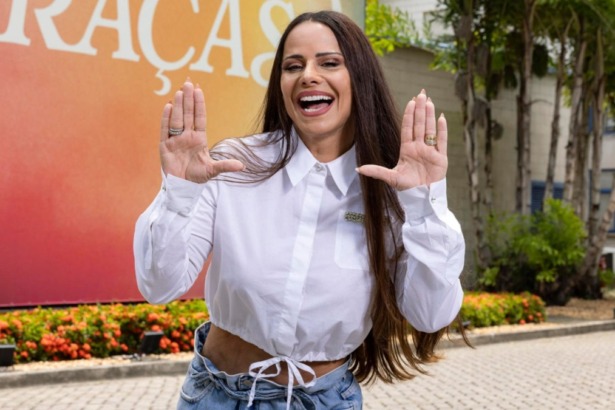 Atriz Viviane Araújo sorrindo e gesticulando em frente a um banner com o título da novela Três Graças, vestindo camisa branca e jeans