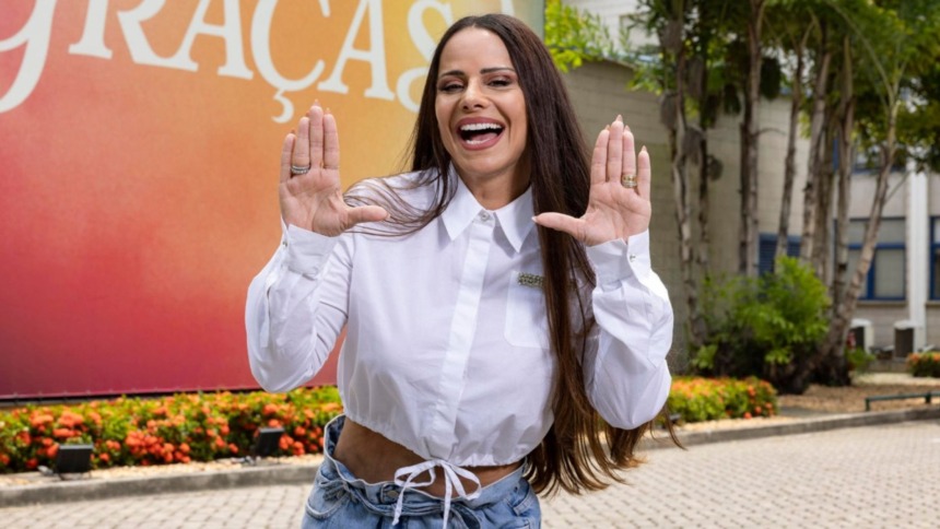 Atriz Viviane Araújo sorrindo e gesticulando em frente a um banner com o título da novela Três Graças, vestindo camisa branca e jeans