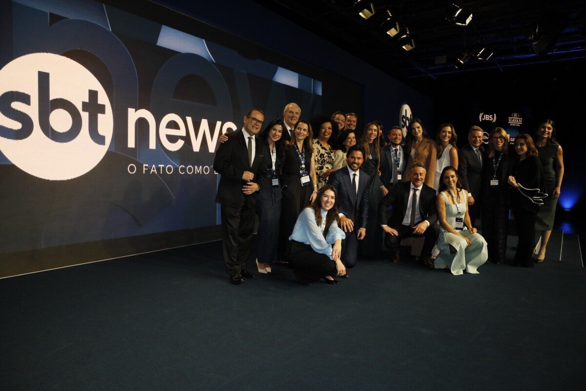 Elenco de apresentadores, jornalistas e diretores do SBT News posam no palco juntos, em frente o telão