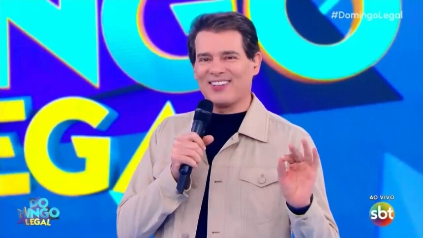 Celso Portiolli apresentando o Domingo Legal em frente ao telão com o logo do programa
