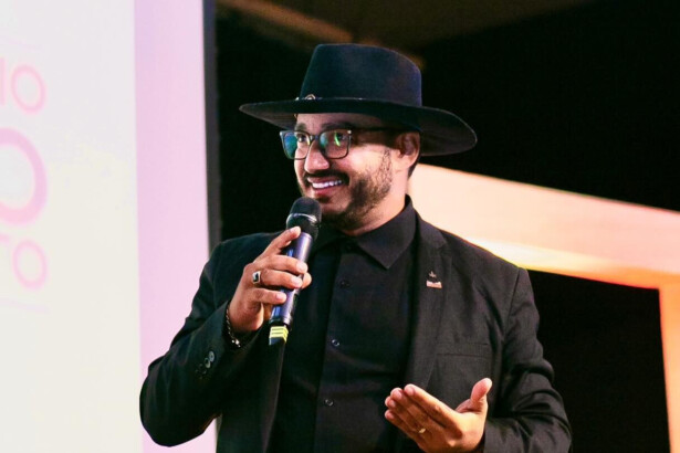 Dudu Morais segura microfone e discursa sorrindo com um chapéu preto na cabeça