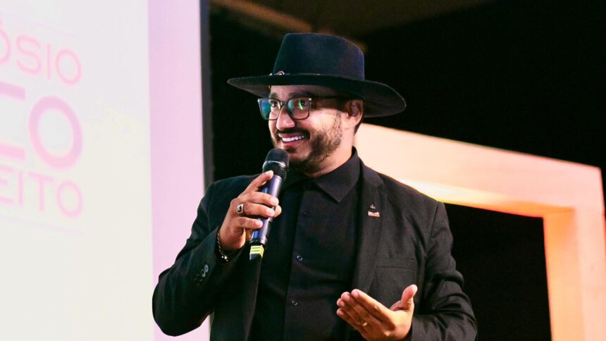 Dudu Morais segura microfone e discursa sorrindo com um chapéu preto na cabeça