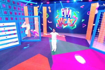Lucas Guimarães veste branco e abre os braços no centro do palco do programa Eita, Lucas!