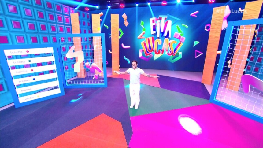Lucas Guimarães veste branco e abre os braços no centro do palco do programa Eita, Lucas!