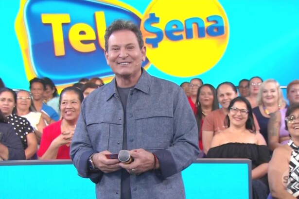 Luis Ricardo sorrir e segura bilhete da Tele Sena Semanal edição 84. Ao fundo um telão com o logo da Tele Sena