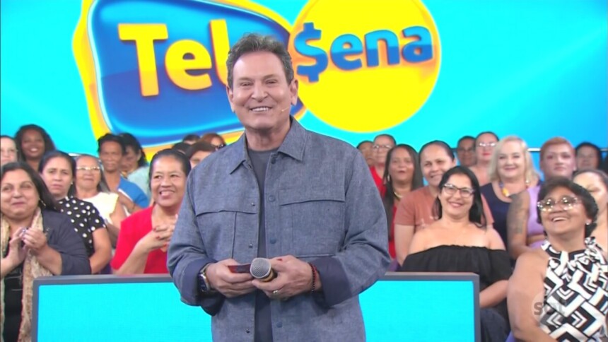 Luis Ricardo sorrir e segura bilhete da Tele Sena Semanal edição 84. Ao fundo um telão com o logo da Tele Sena