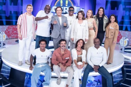 Tom Cavalcante com um terno cinza claro posa com os 11 participantes do reality show "Acerte ou Caia!" no palco, todos sorrindo e em clima de festa.