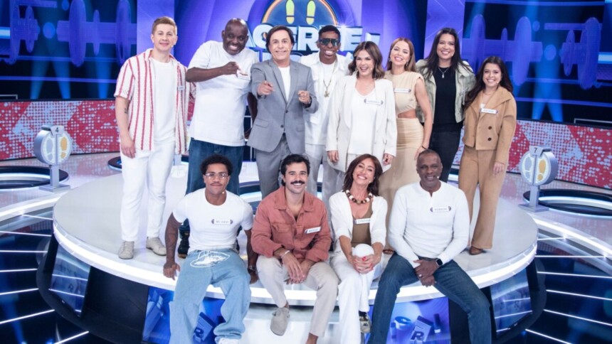 Tom Cavalcante com um terno cinza claro posa com os 11 participantes do reality show "Acerte ou Caia!" no palco, todos sorrindo e em clima de festa.