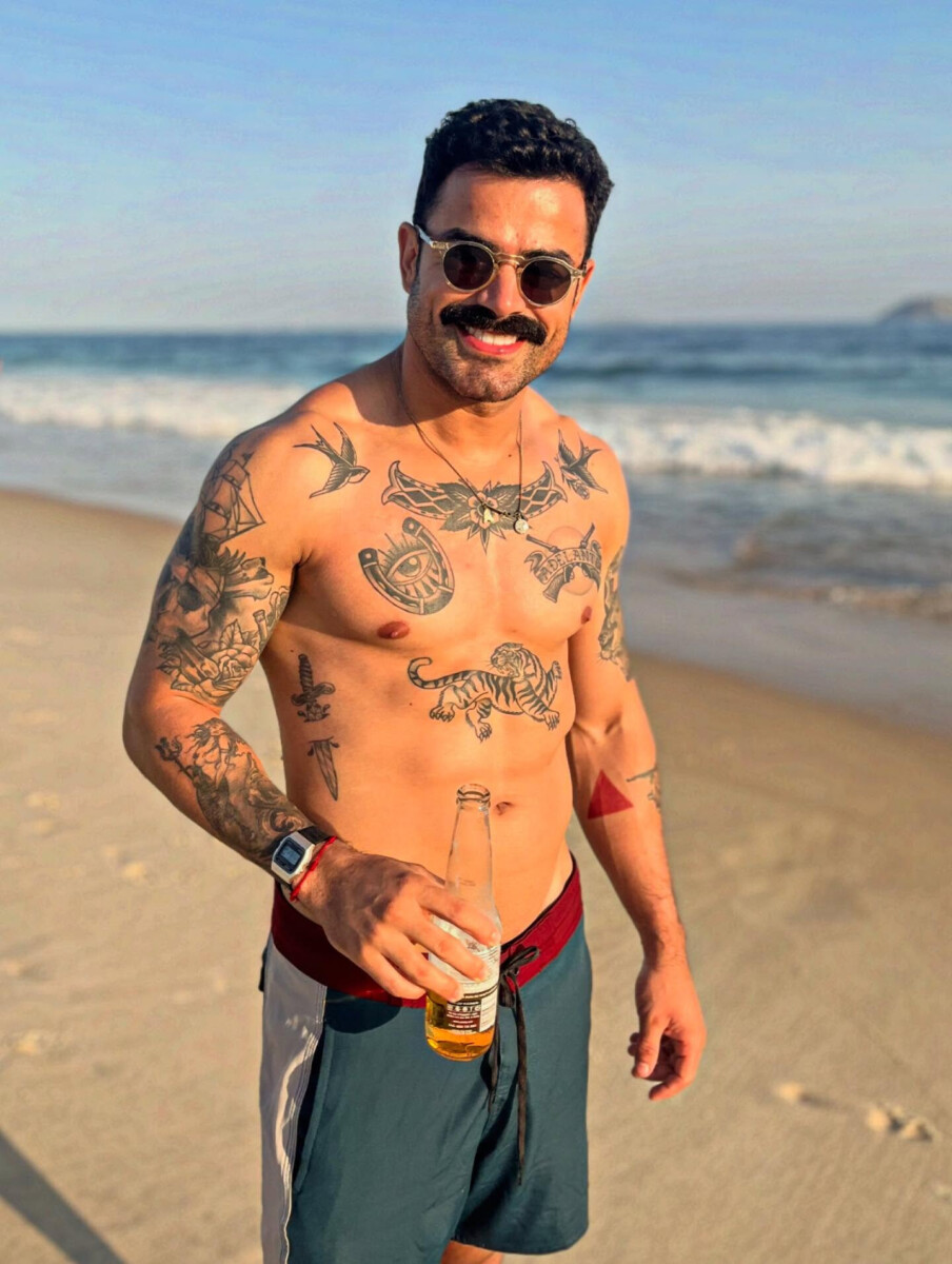 Repórter Túlio Amancio sem camisa, mostrando corpo malhado e tatuado. Ele está em uma praia segurand uma cerveja e sorridente