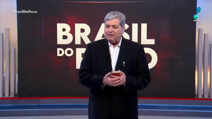 Datena segura telefone celular nas mãos em frente ao telão com o logo do Brasil do Povo no cenário da RedeTV!