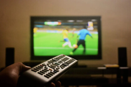 Controle remoto em frente a uma tela de TV com jogadores em uma transmissão como da Cazé TV