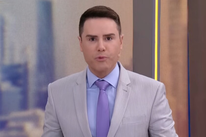 Luiz Bacci de terno e grava fazendo bico com a boca no cenário do SBT