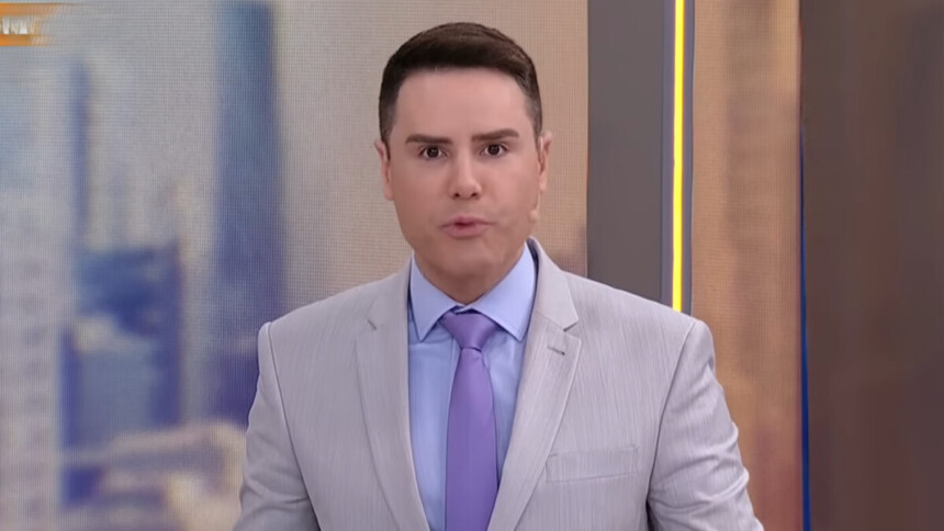 Luiz Bacci de terno e grava fazendo bico com a boca no cenário do SBT
